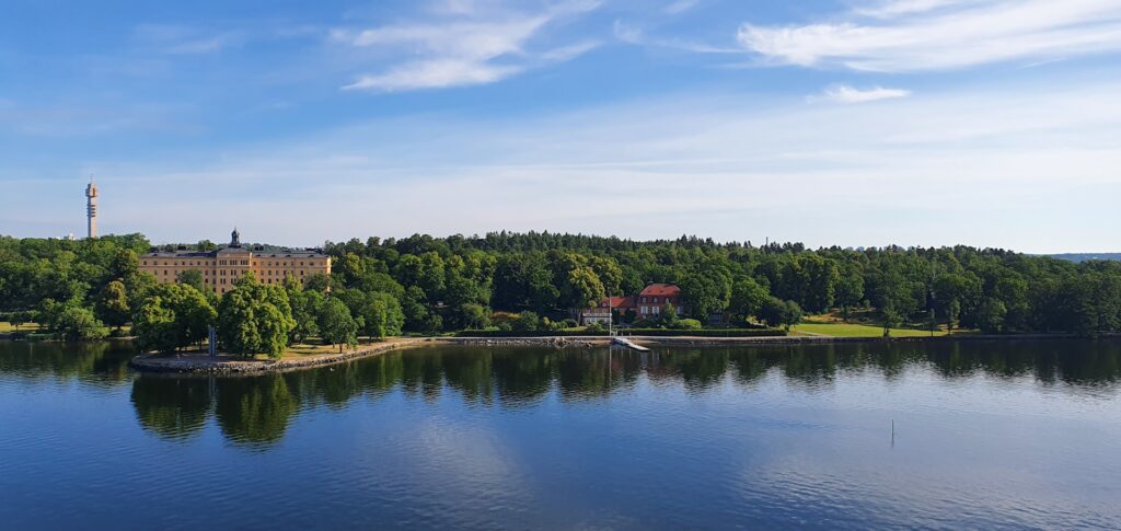 Paisagem natural em Estocolmo com lago calmo, área verde arborizada e céu azul. Foto: Kabir Baidhya / Unsplash.