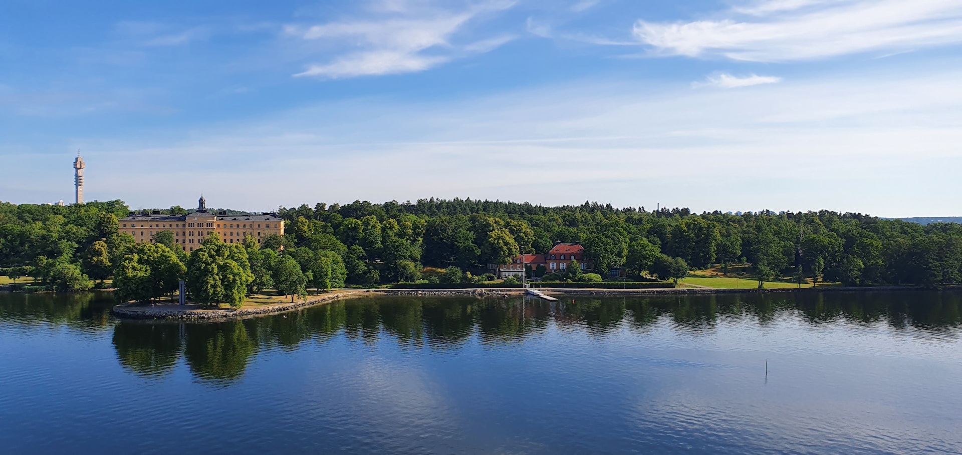 Paisagem natural em Estocolmo com lago calmo, área verde arborizada e céu azul. Foto: Kabir Baidhya / Unsplash.